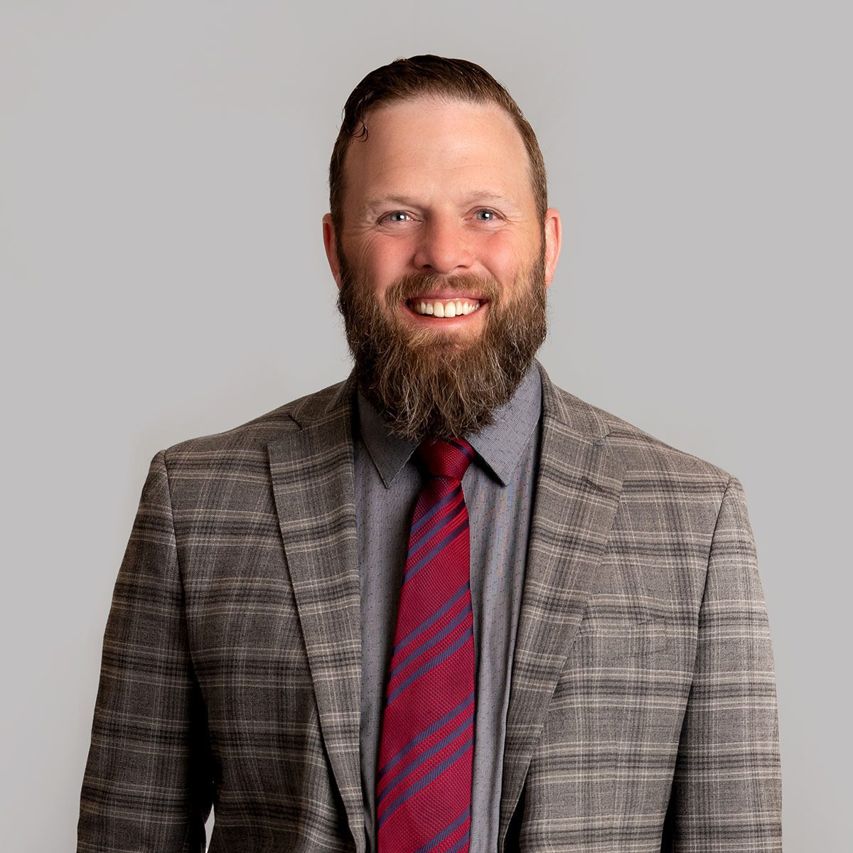 A person with short brown hair and brown beard smiles at the camera. He's wearing a grey plaid jacket, a grey shirt, and a red tie with blue stripes.
