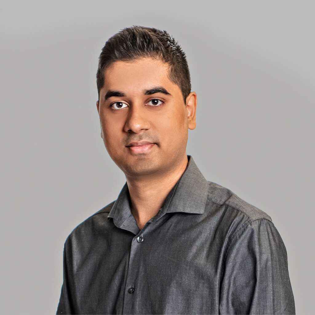 Amir A person with short dark brown hair looks at the camera. They are wearing a dark grey buttoned up shirt.