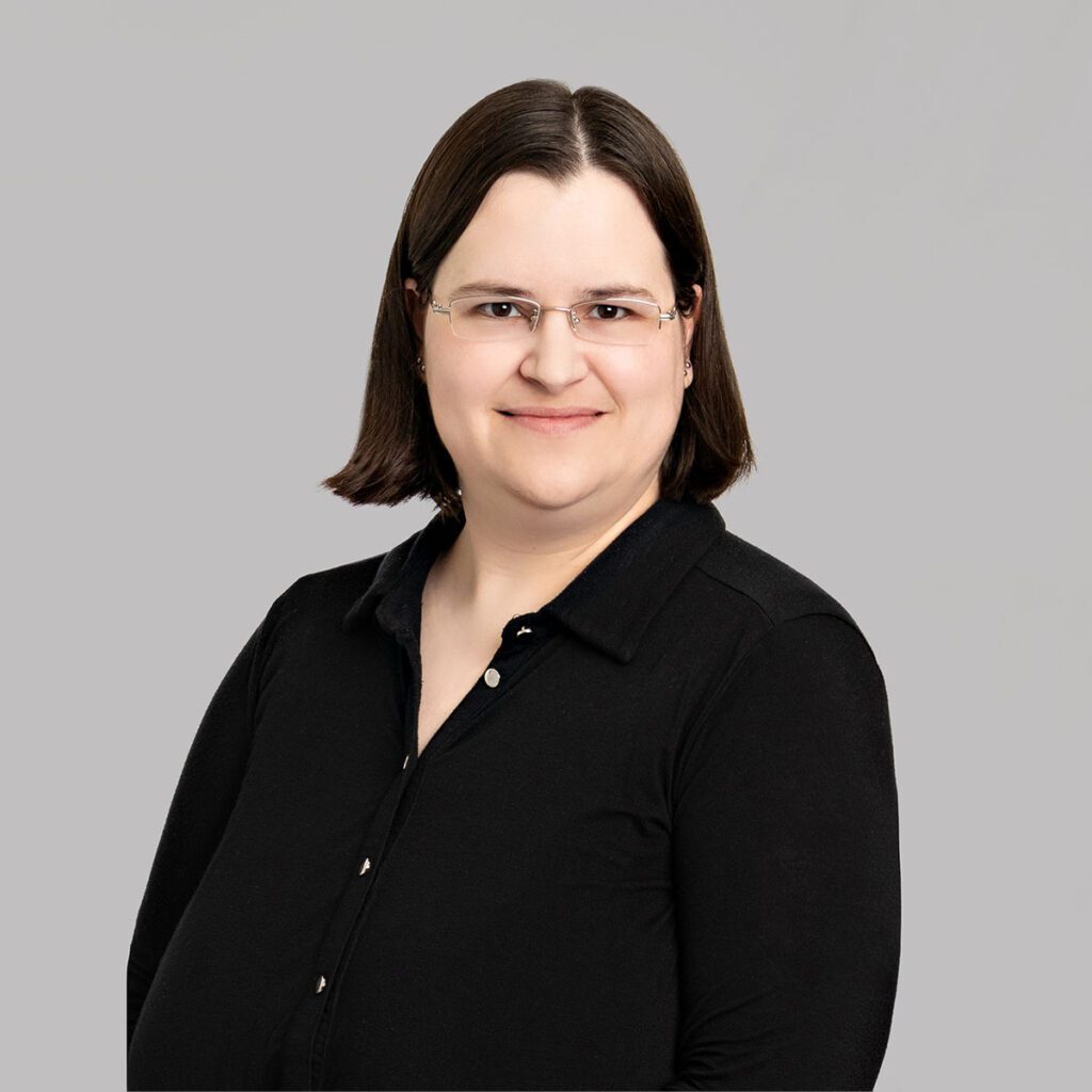 A person with short brown hair smiles at the camera. They are wearing a black buttoned up shirt.