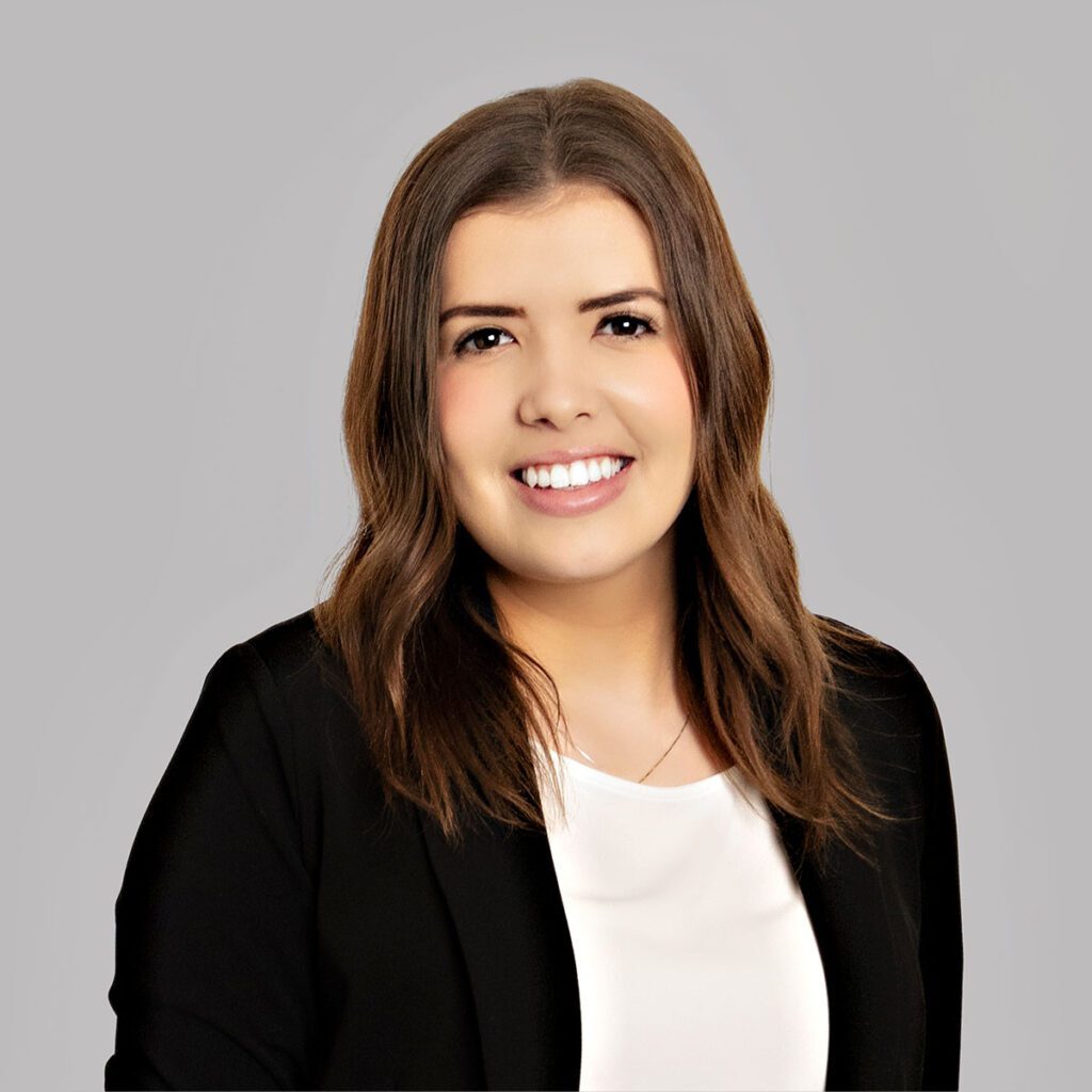 Kayla A person with brown hair smiles at the camera. They are wearing a black blazer with a white blouse underneath.