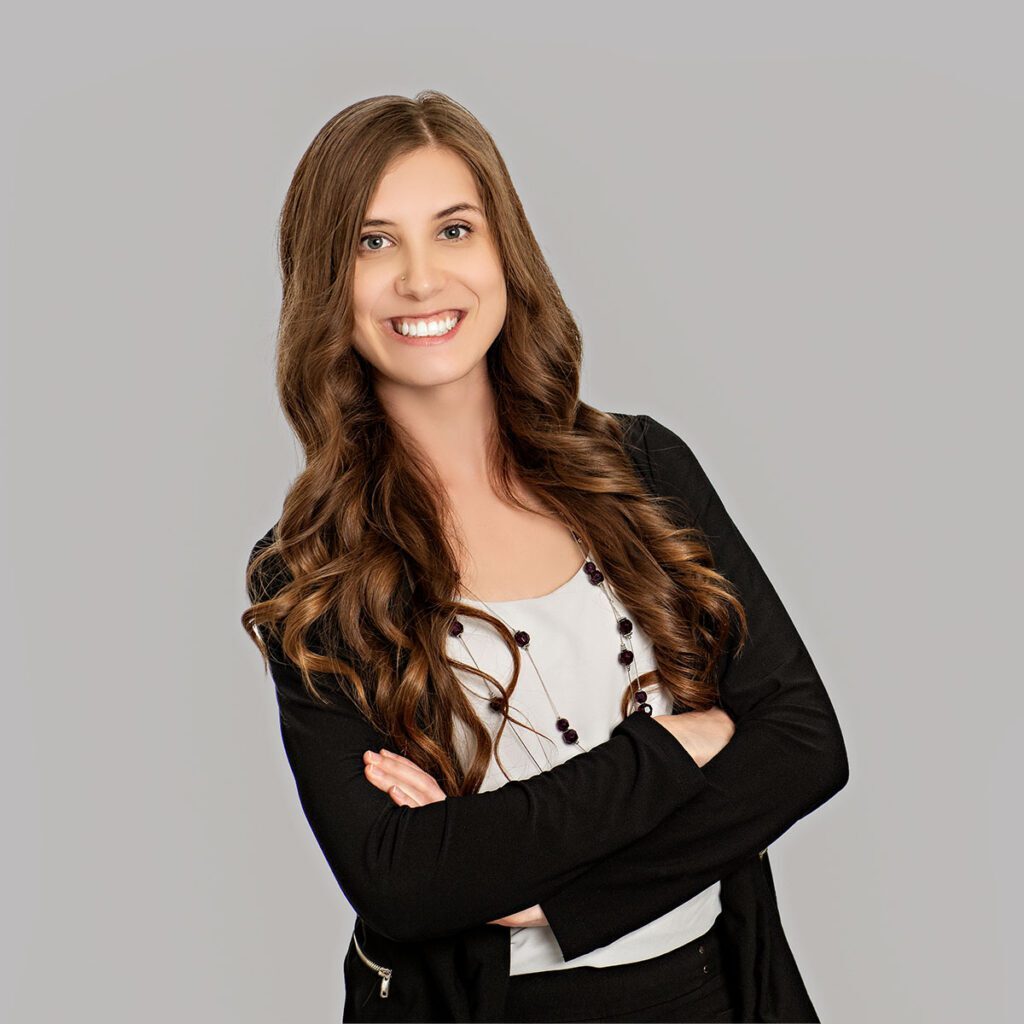 A person with very long brown hair smiles at the camera. They are wearing a black blazer with a white blouse underneath, and a long gold necklace accented wth black beads.