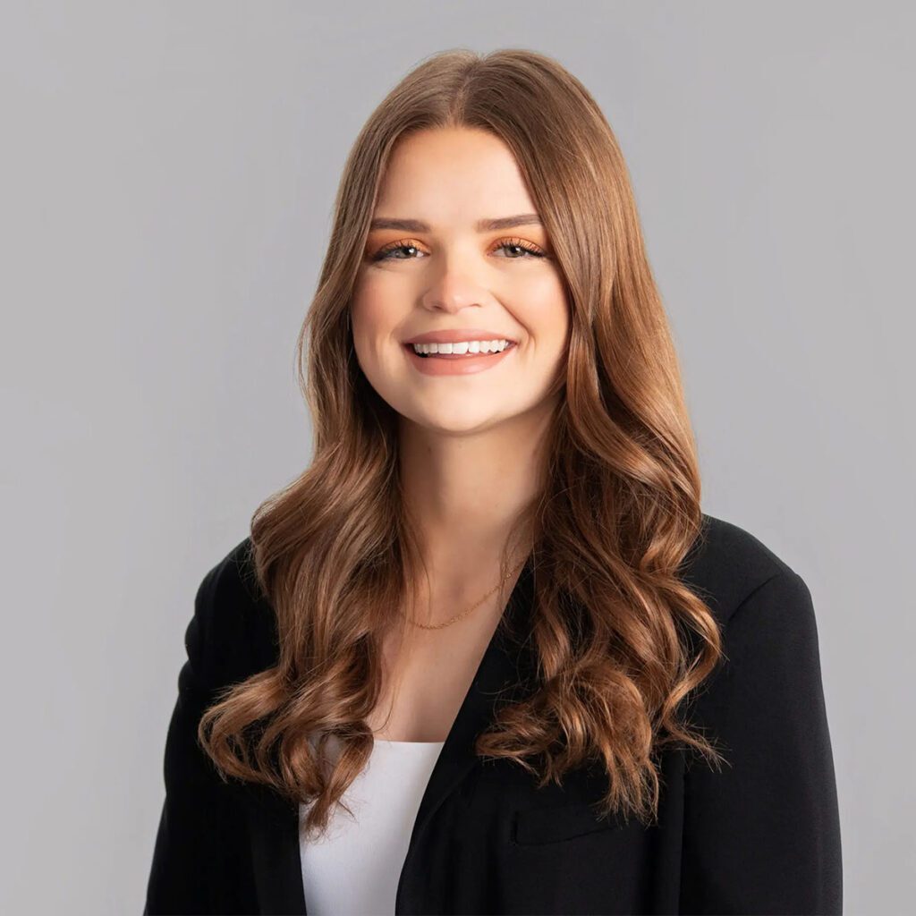 A person with long light brown hair smiles at the camera. They are wearing a black blazer with a white blouse underneath.