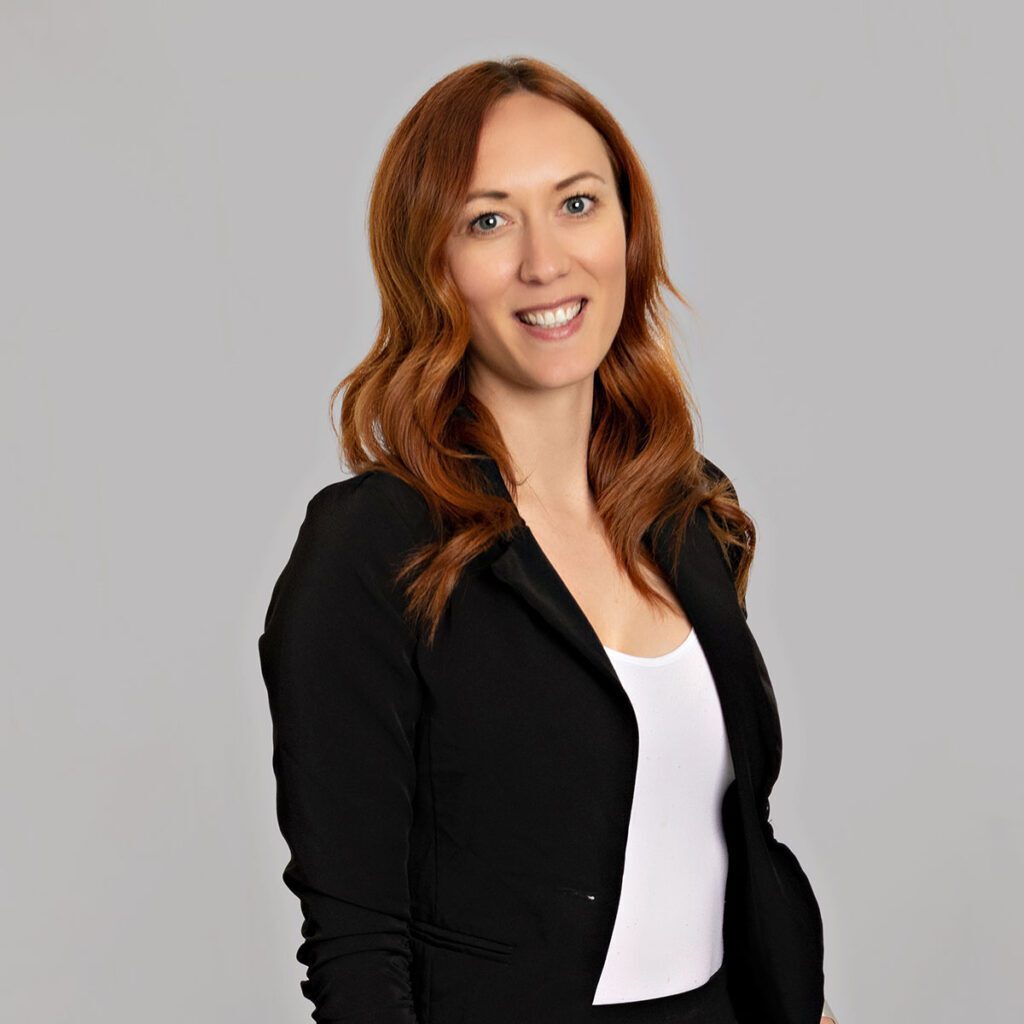 A person with long red hair smiles at the camera. They are wearing a black blazer with a white blouse underneath.